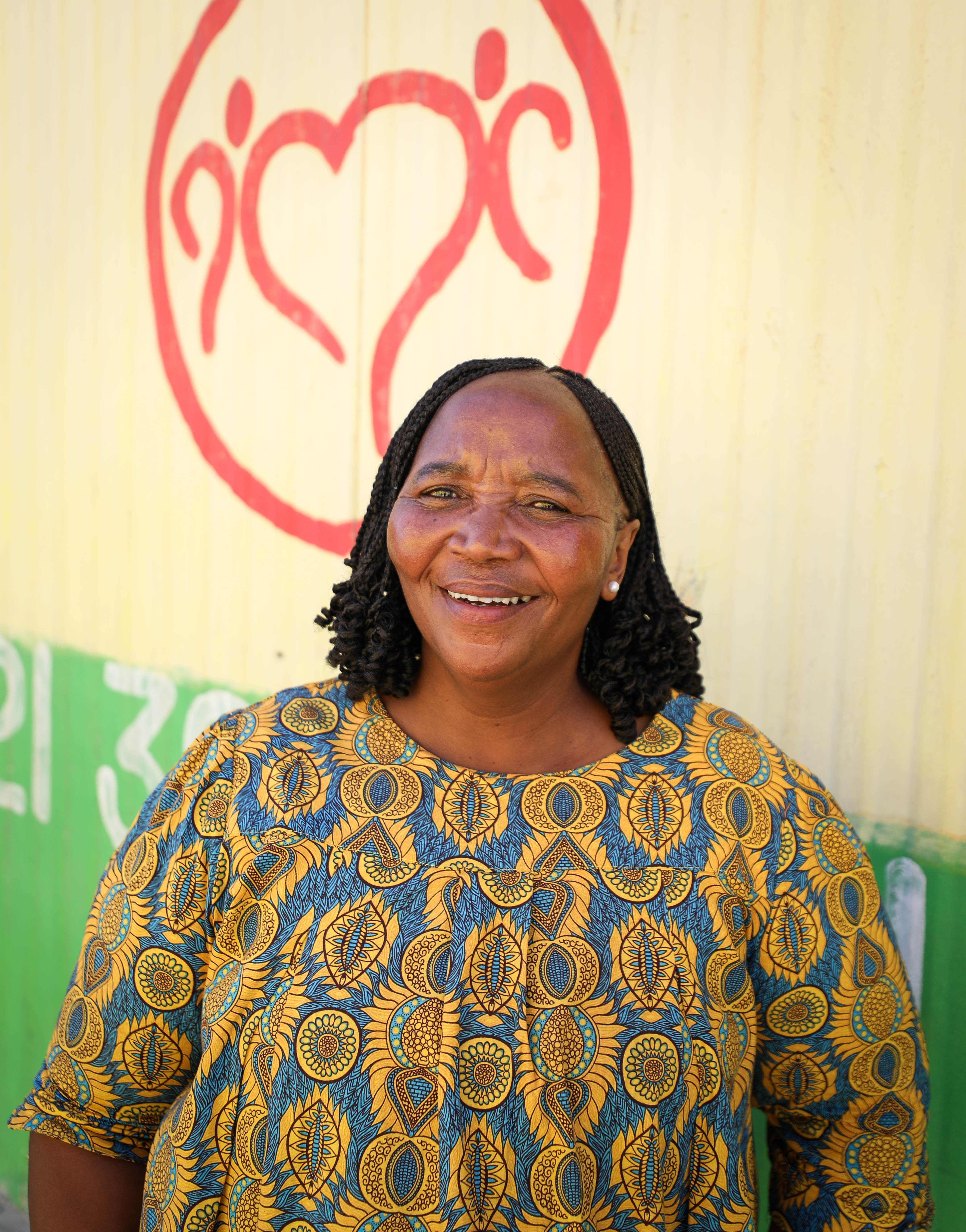 A woman with black braids stands in front of a yellow building smiling.