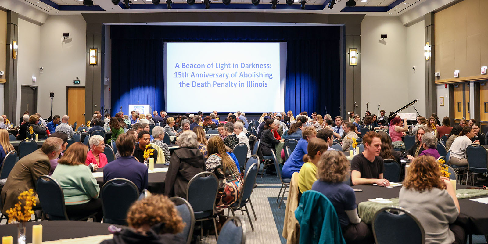 100 people sit in round tables of ten in a large room.